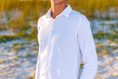 Young man in a white shirt and light pants standing on the beach, with golden seagrass and a vibrant sky in the background
