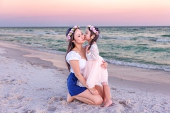 Mother sitting on the sand, holding her daughter close and kissing her cheek during a beach sunset