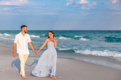 Couple posing affectionately on the beach, wearing casual yet sophisticated outfits, with soft waves and blue skies in the background