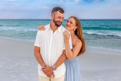 Smiling couple holding hands and walking along the beach, dressed in white and blue outfits, with waves lapping at the shore