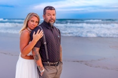 Couple standing close on a sandy beach, with the ocean waves and a vibrant blue sky behind them